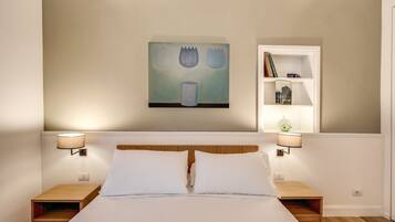 A neatly made bed with two pillows, a wall-mounted painting, a bookshelf with books and decorative items, and two bedside lamps.
