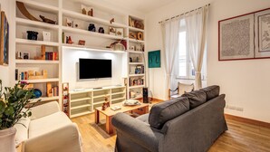 A living room with a flat-screen TV, a grey sofa, a coffee table, and a bookshelf filled with books and decorative items. - Dopodomani Suite (Rome)