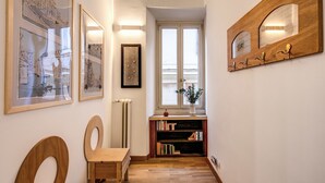 A hallway with framed artwork, a wooden chair, a bookshelf, and a window with a view. - Dopodomani Suite (Rome)