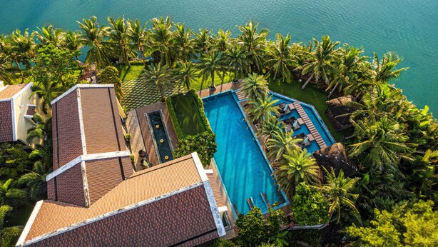 An aerial view of a resort with a large swimming pool surrounded by palm trees and a clear view of the ocean. - Koi Resort and Spa Hoi An (Da Nang)