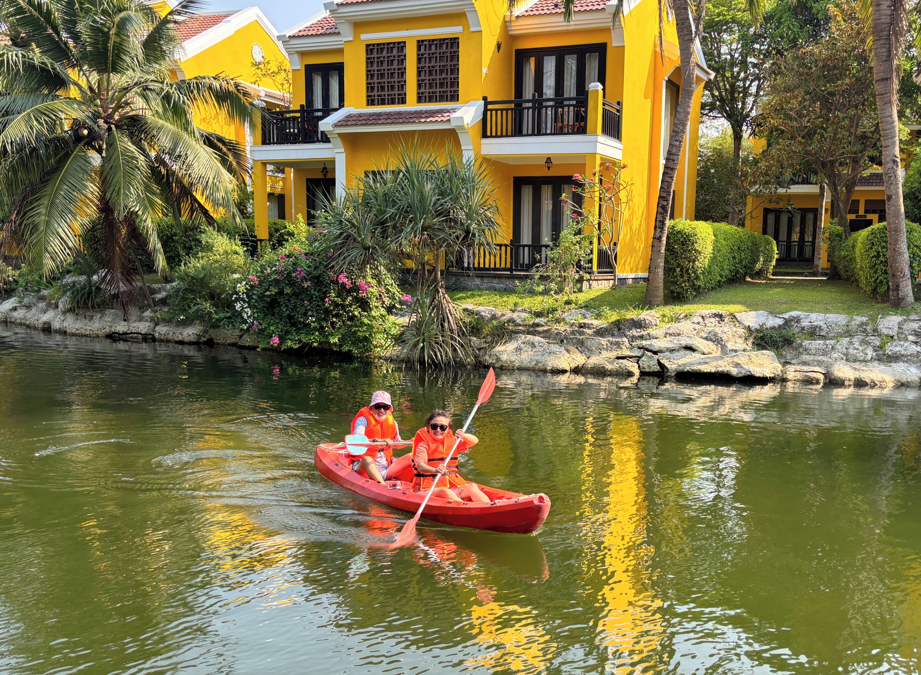 A yellow building with a balcony, a water body, and a red boat with two people.