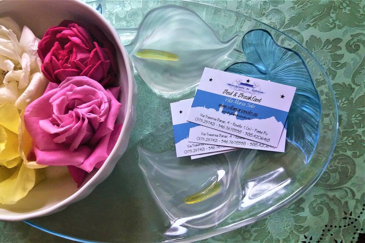 A bowl of artificial flowers in yellow, white, and pink, and a clear glass dish with two business cards on a patterned tablecloth.