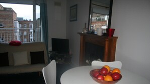 Un área de comedor con una mesa redonda, un cuenco de fruta, un sofá blanco y vistas de edificios a través de una ventana.