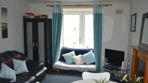 Una sala de estar con un sofá de cuero negro, una silla blanca, una mesita redonda con una planta y un televisor sobre un mueble.