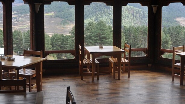 A wooden dining area with tables and chairs, large windows offering a view of a forested landscape. - Dewachen Resort (Paro)