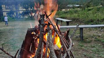 Una fogata con leños y ramas, rodeada por un área de césped con un banco de madera y una persona de pie al fondo.
