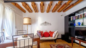 A living room with a wooden ceiling, a sofa with red cushions, a TV stand with books, and a dining table with chairs.