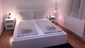 A double bed with floral bedding, two bedside tables with lamps, a nightstand, a framed picture on the wall, and a window with blinds. - Toldi Apartments (Pecs)
