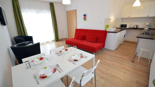 A modern apartment with a dining area, a red sofa, a kitchen, and a white table set for two. - Toldi Apartments (Pecs)