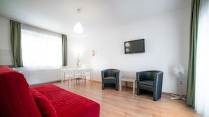 A modern room with a red bed, a TV, two chairs, a small table, and a standing fan. - Toldi Apartments (Pecs)