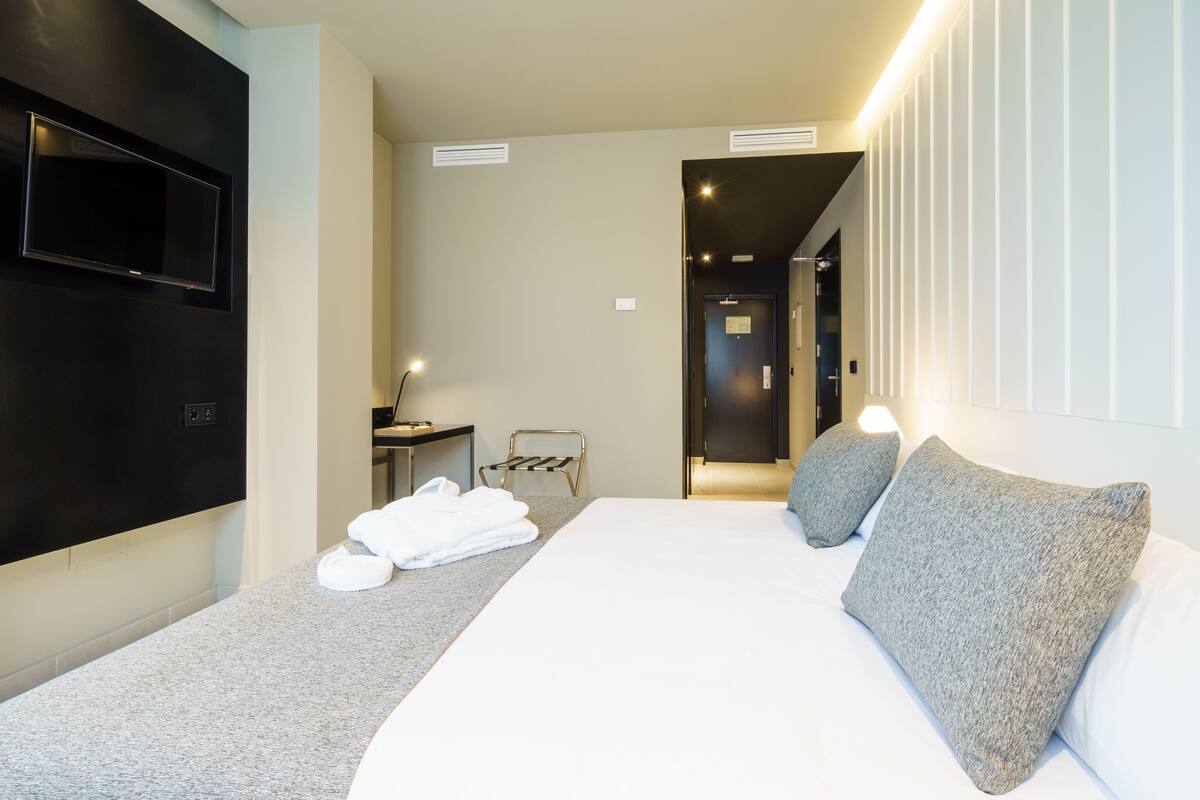 A modern hotel room with a large bed, a flat-screen TV mounted on the wall, a desk with a chair, and a small table with a lamp.