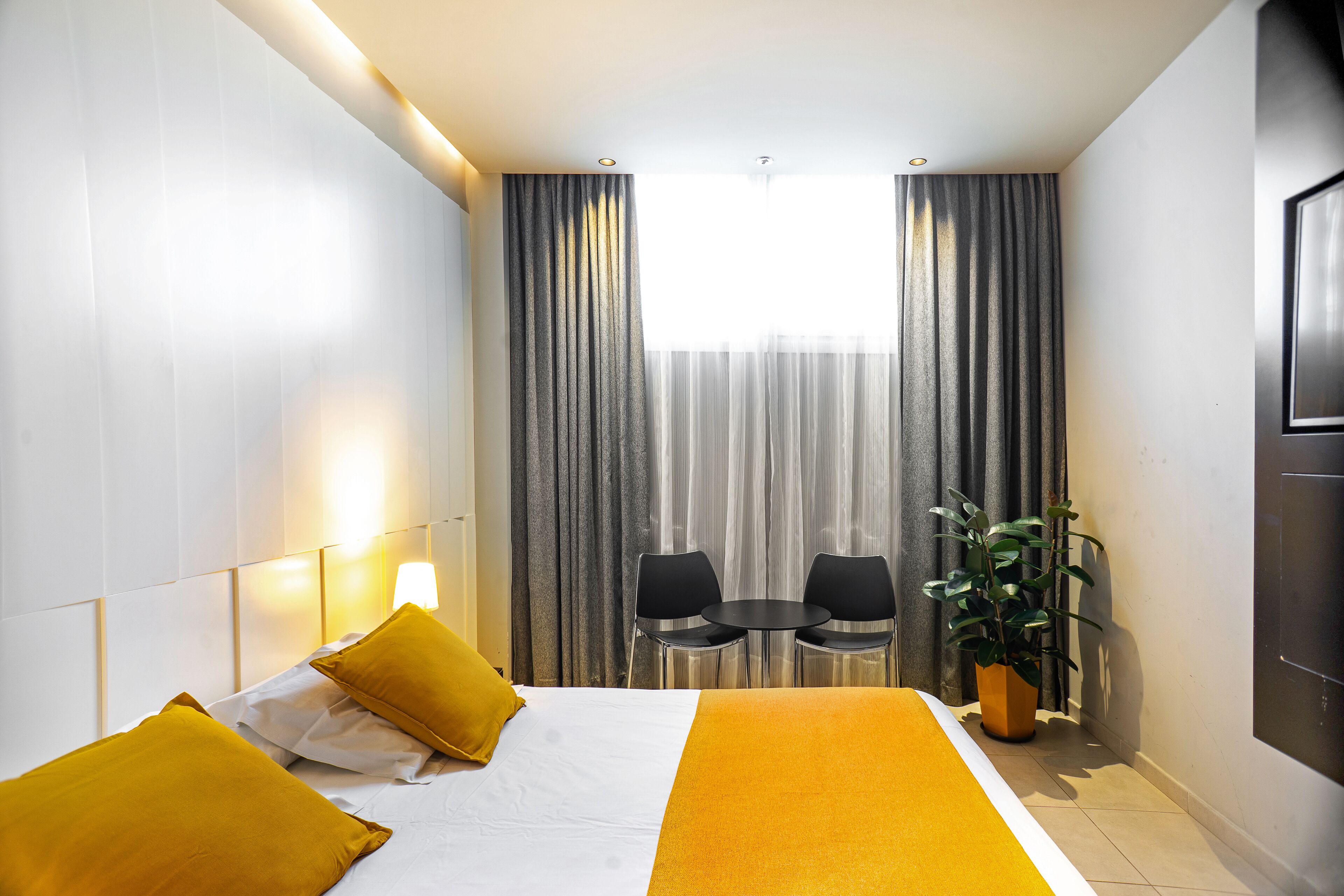 A hotel room with a bed, two yellow pillows, a bedside table, a potted plant, and a window with curtains.