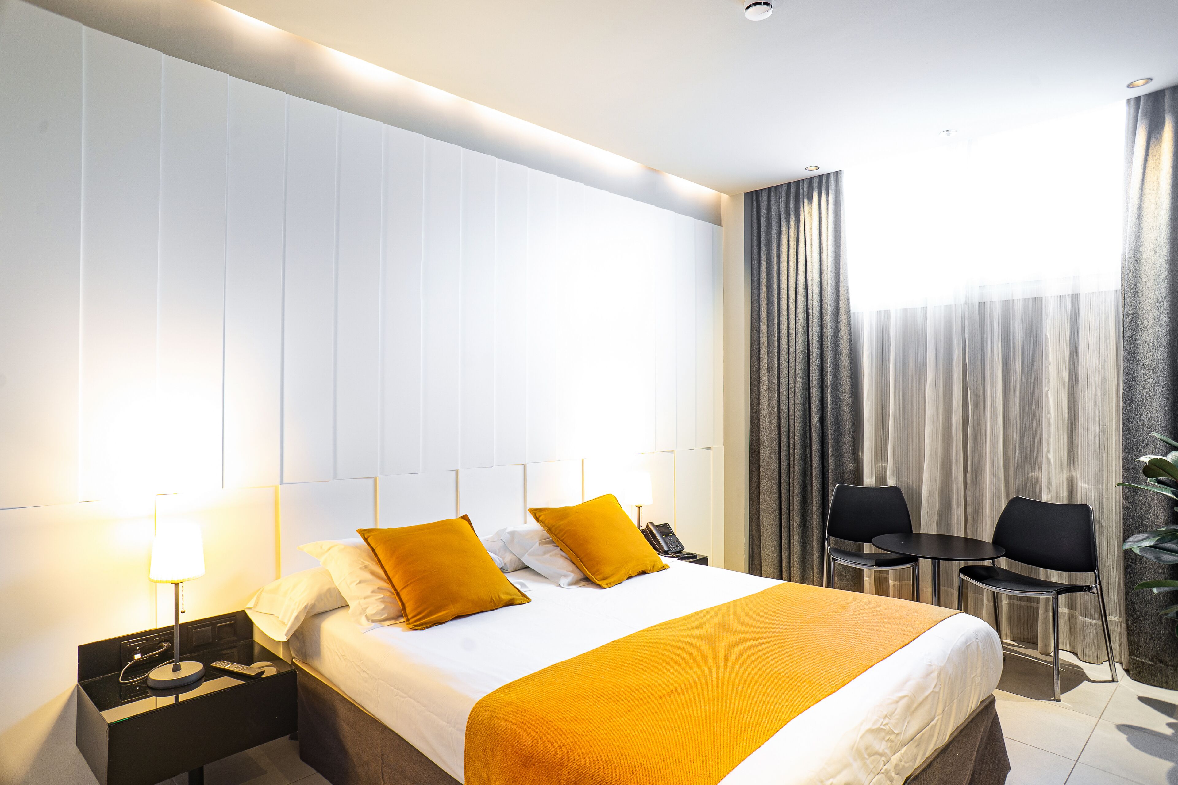 Een hotelkamer met een bed, nachtkastje, lamp en een klein tafeltje met stoelen.