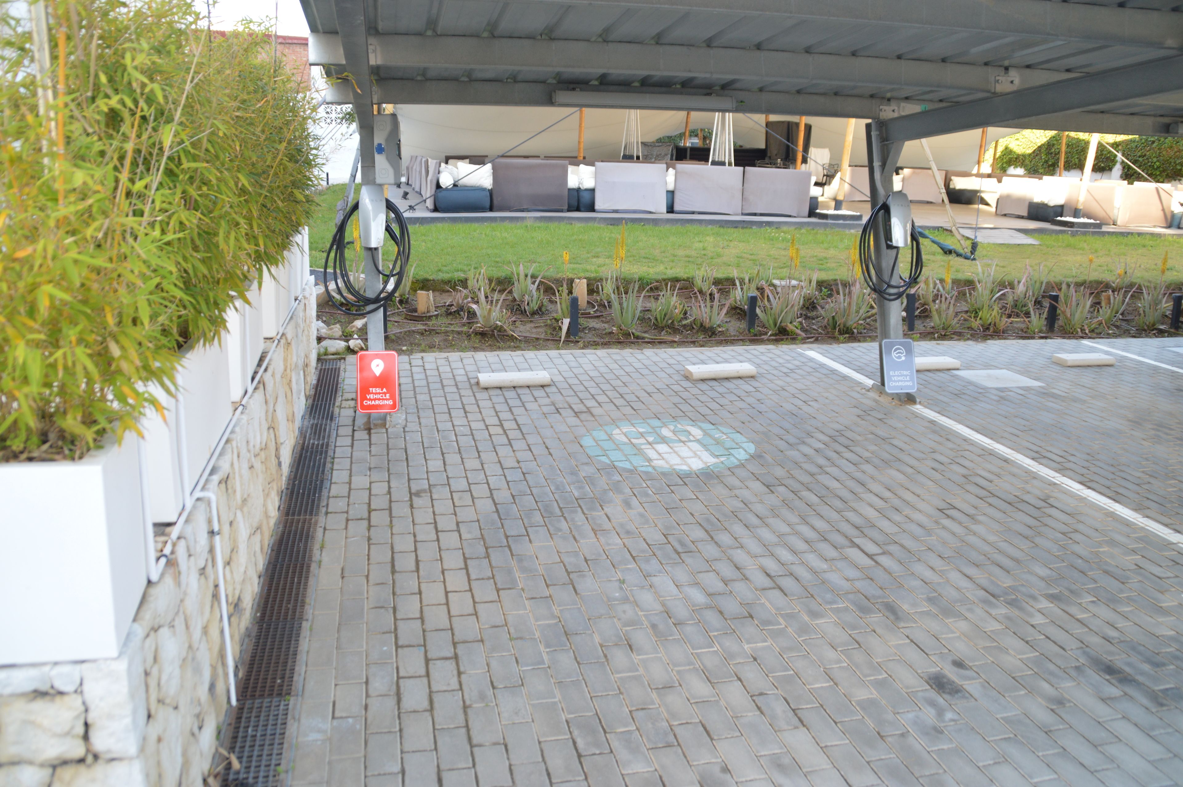 A parking area with a Tesla charging station, a covered seating area, and a grassy patch.