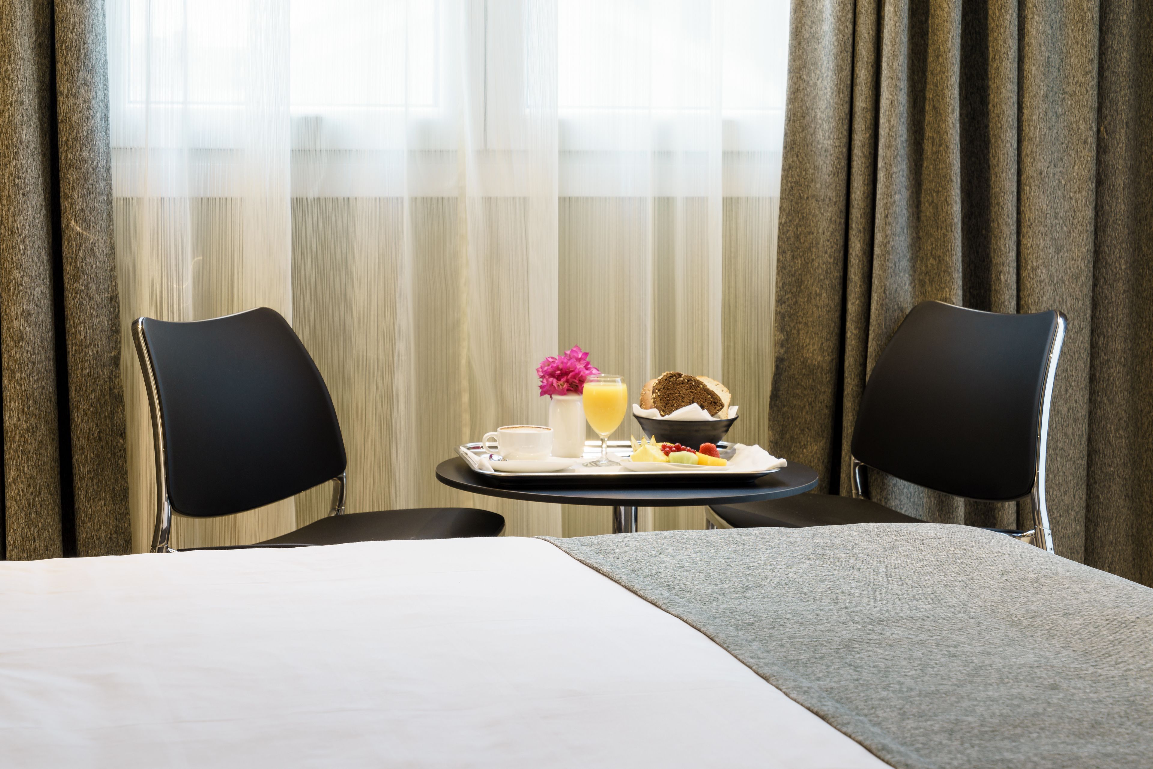 Une chambre d’hôtel avec un lit, deux chaises noires, une petite table ronde avec des articles pour le petit-déjeuner et des rideaux légers.
