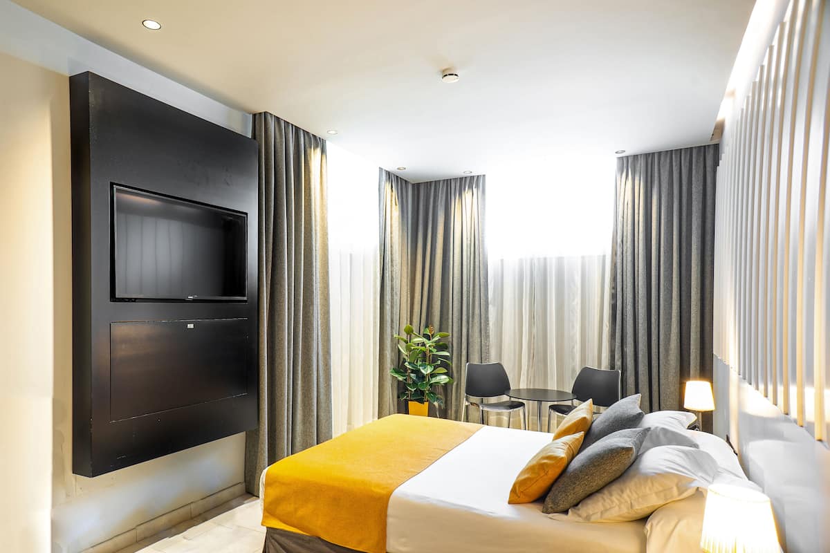 A modern hotel room with a large bed, a flat-screen TV mounted on the wall, a small table with chairs, and a potted plant.