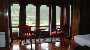 Une pièce avec un plancher en bois, deux chaises rouges, une petite table et un lit. De grandes fenêtres offrent une vue sur un paysage verdoyant.