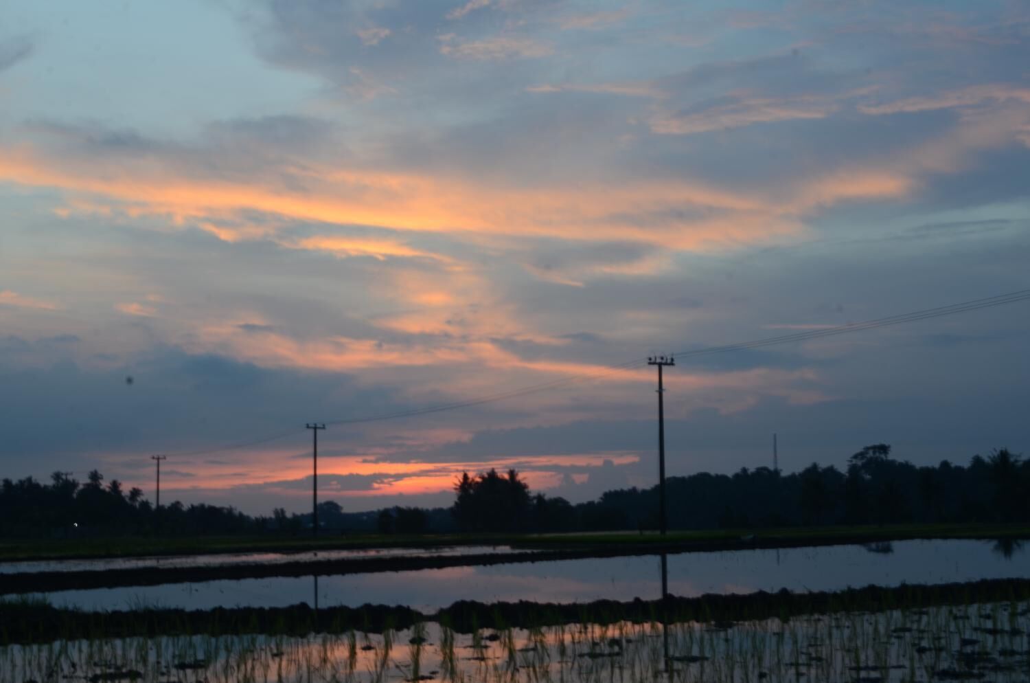 A sunset over a rural landscape with power lines and a calm body of water reflecting the sky.