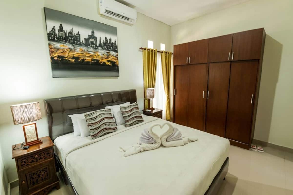 A hotel room with a bed, a wardrobe, a painting on the wall, and a nightstand with a lamp.