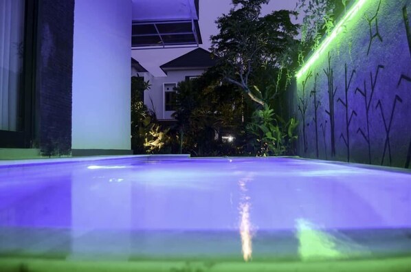 An indoor swimming pool with a clear water surface, surrounded by a modern building with large windows and a garden area with trees and plants. - Beluran Serene Guesthouse (Tabanan)
