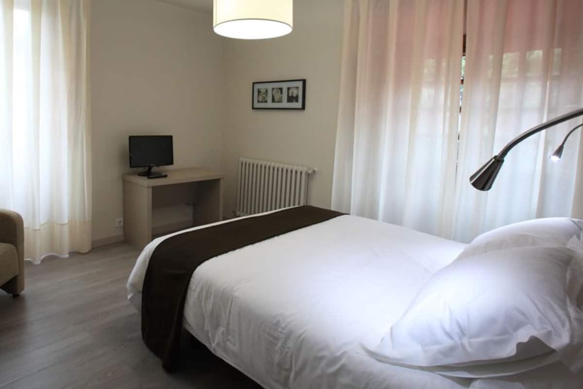 A hotel room with a bed, a chair, a desk with a computer, and a window with curtains.