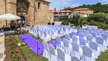 An outdoor wedding venue with rows of white chairs and purple tablecloths, stone architecture, and a garden setting.