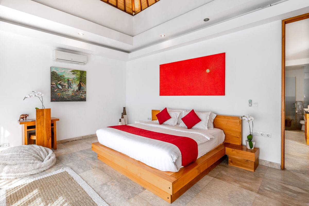 A modern bedroom with a large bed, a red accent wall, and a painting of a tropical scene.