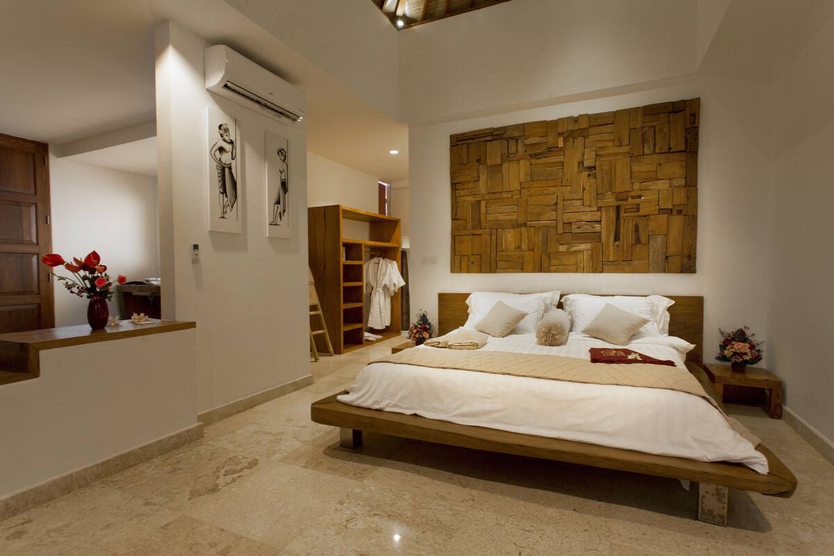 A spacious bedroom with a large bed, a wooden headboard, a wall-mounted wooden panel, a small bedside table, and a nightstand.
