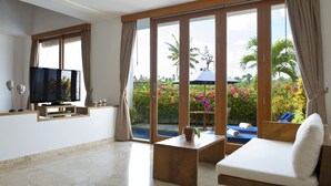 A modern living room with a flat-screen TV, a sofa, a coffee table, and a large sliding glass door leading to a pool area with a view of tropical plants and a palm tree. - Villa CinCin (Canggu)
