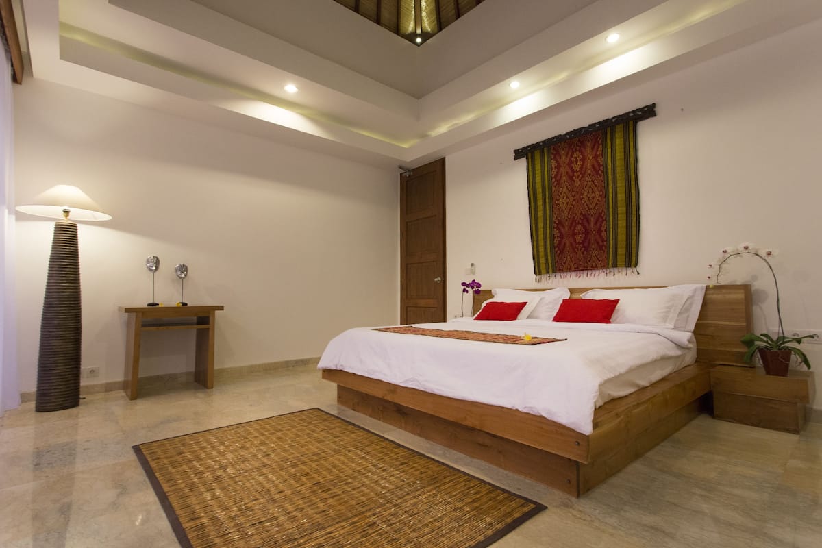 A bedroom with a wooden bed, a nightstand, a lamp, a small table, and a decorative plant.