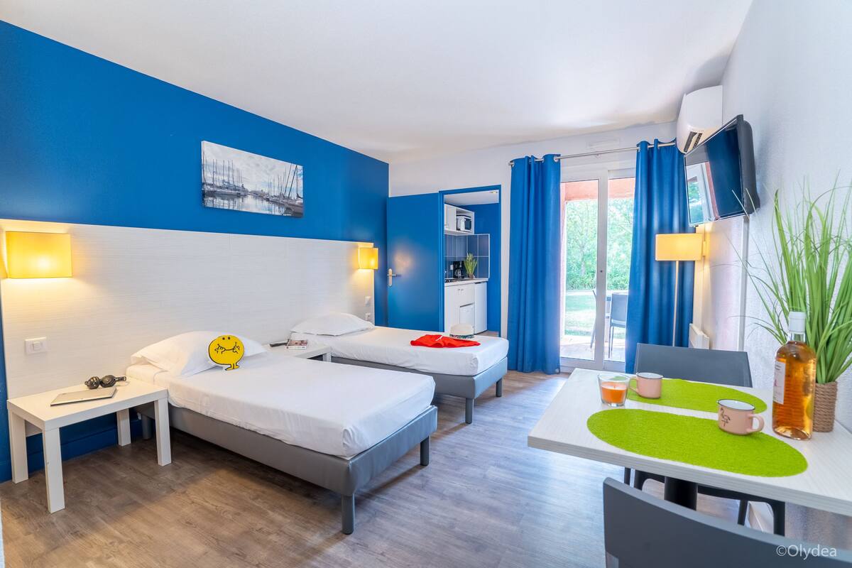 A hotel room with two single beds, a desk, a small table with a green placemat, a TV mounted on the wall, and a window with blue curtains.