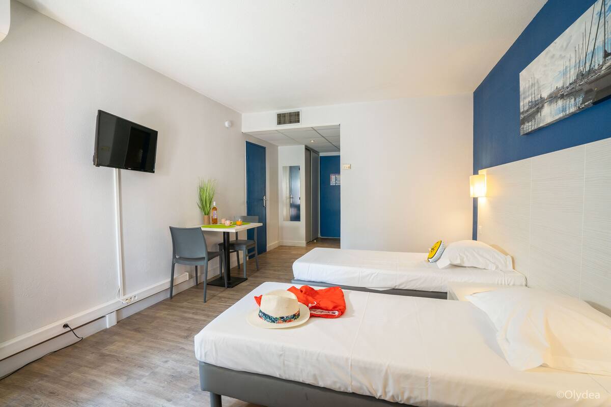 A hotel room with two beds, a desk with a chair, a TV mounted on the wall, and a painting above the bed.