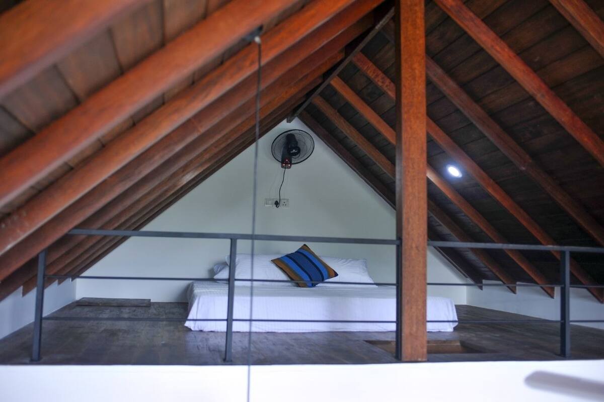Un dormitori al menjador amb sostre inclinat, un llit amb roba de llit blanca i un coixí blau, i un ventilador de sostre.