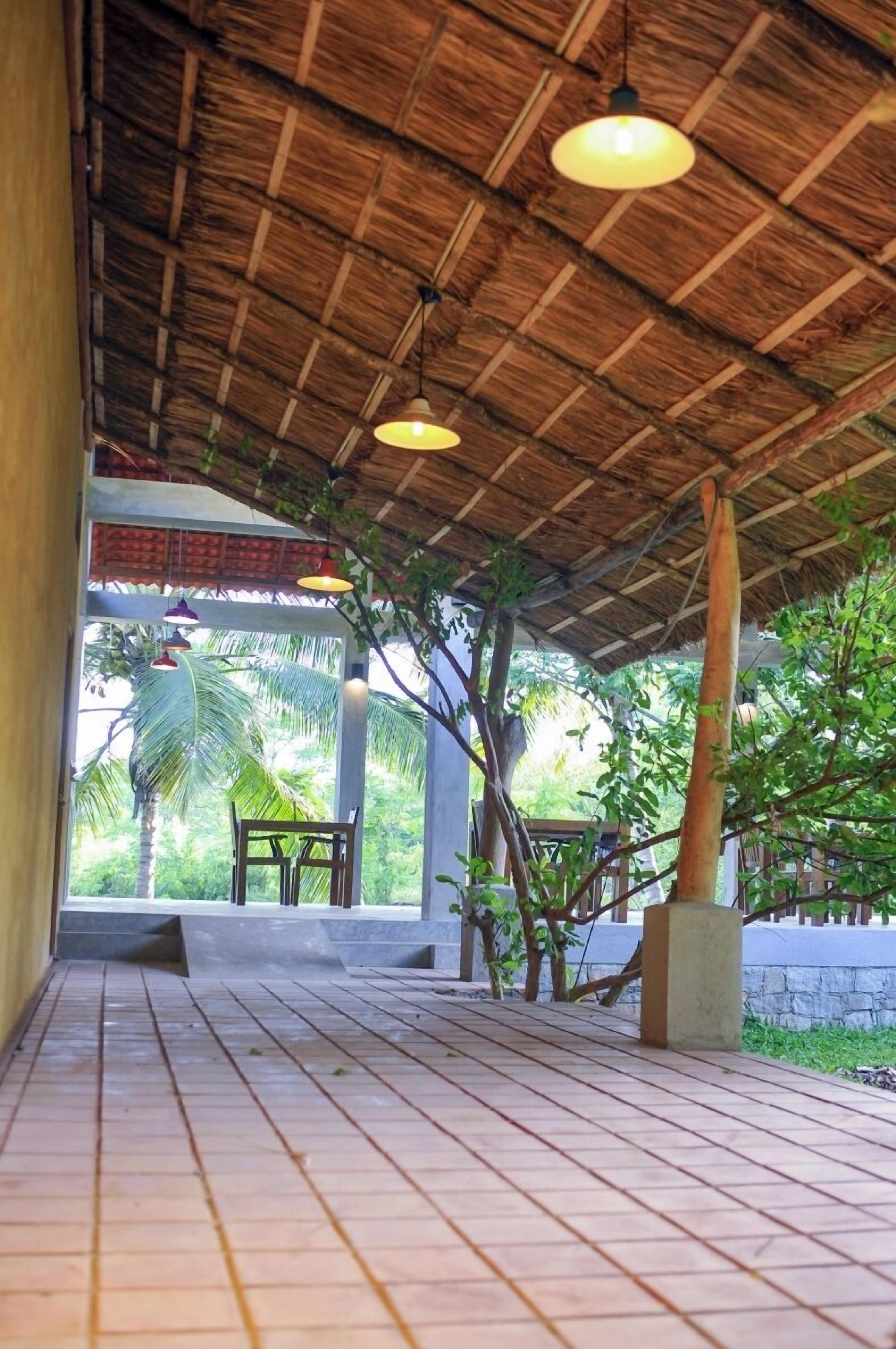 A covered outdoor area with a tiled floor, wooden benches, hanging lights, and a view of greenery.