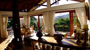 A spacious room with a wooden ceiling, large windows with curtains, a dining table with chairs, and a view of greenery outside. - Lis Bleu Pousada (Tiradentes)