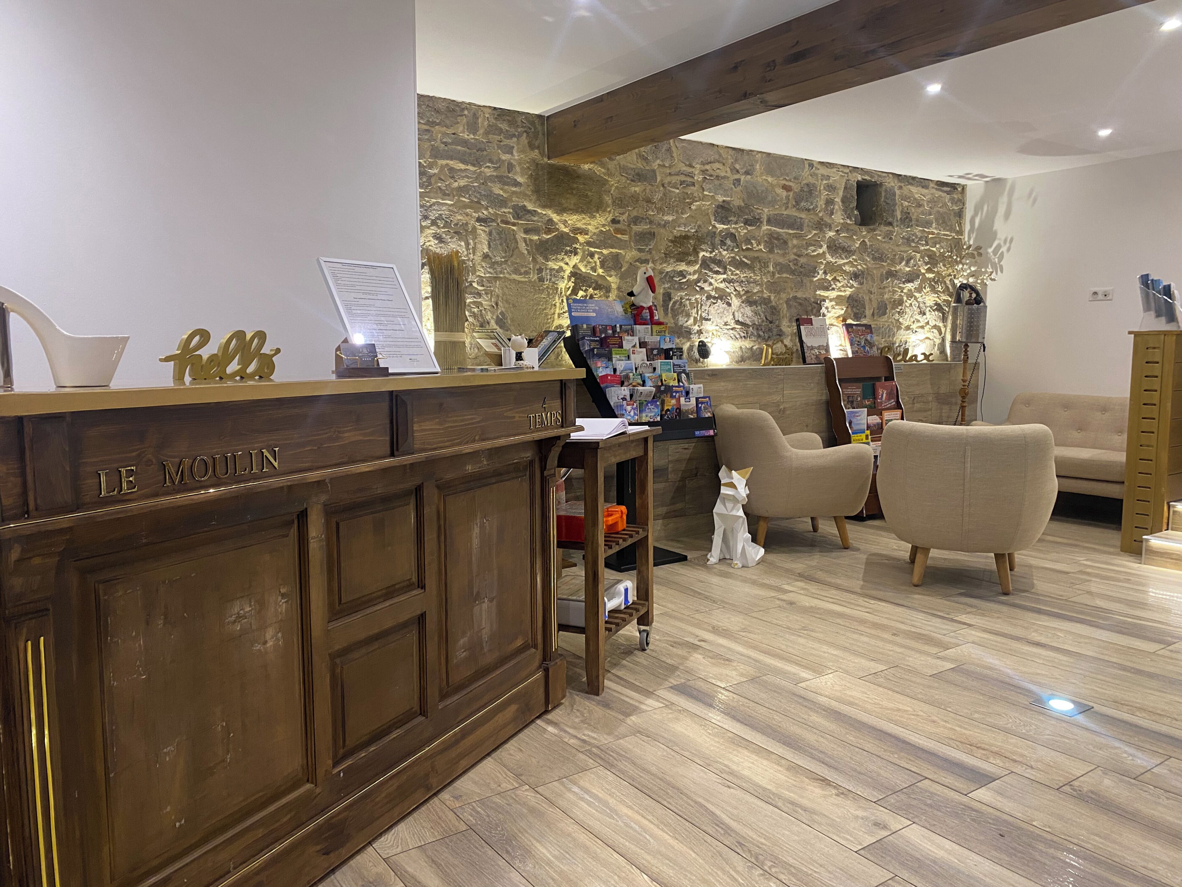 Een receptiebalie met de tekst 'LE MOULIN', een stenen muur, een houten vloer en een zithoek met een bank en stoelen.