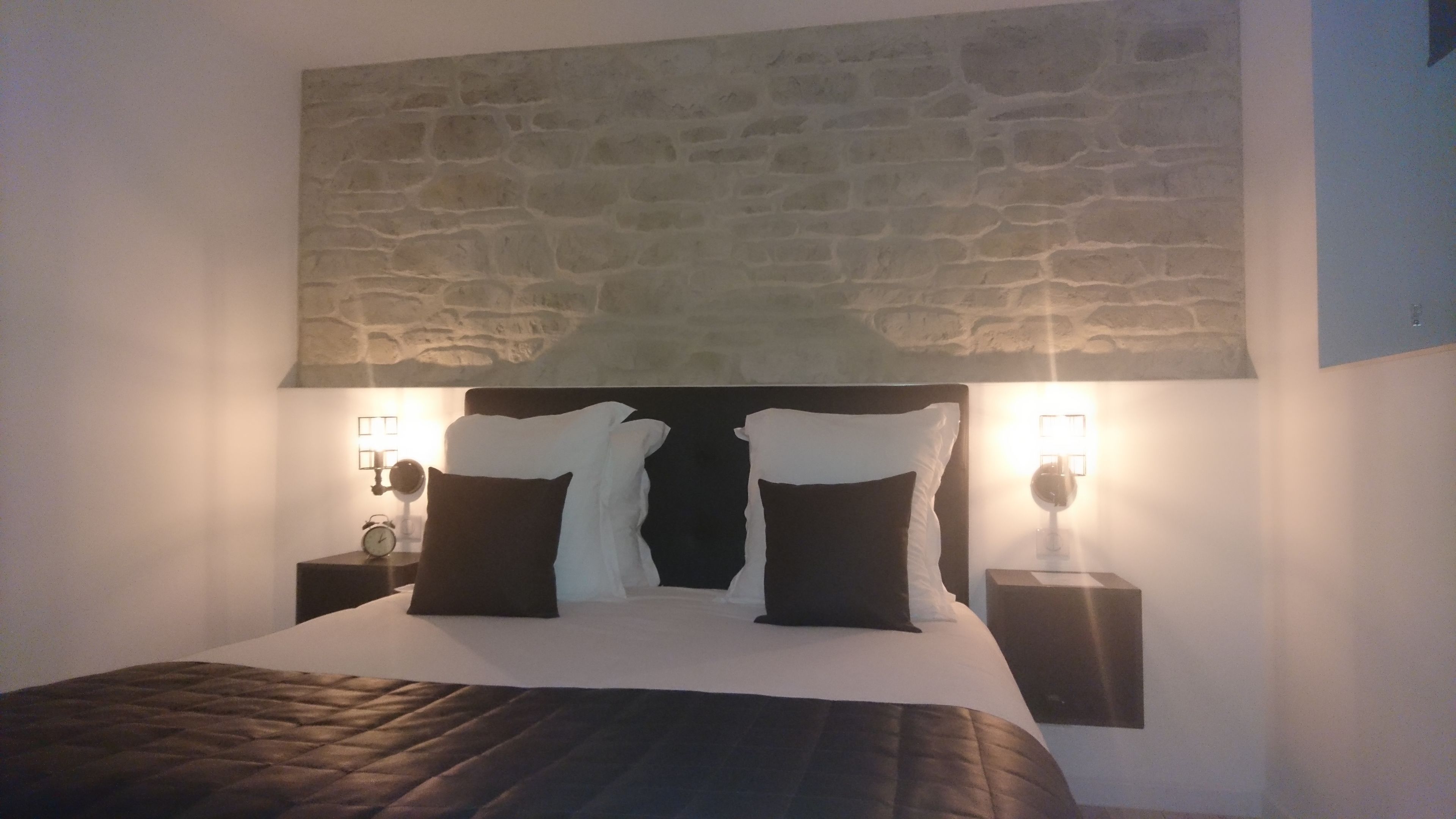 Una cama con cubrecama acolchado, dos mesitas de noche con lámparas y una pared de piedra sobre la cama.