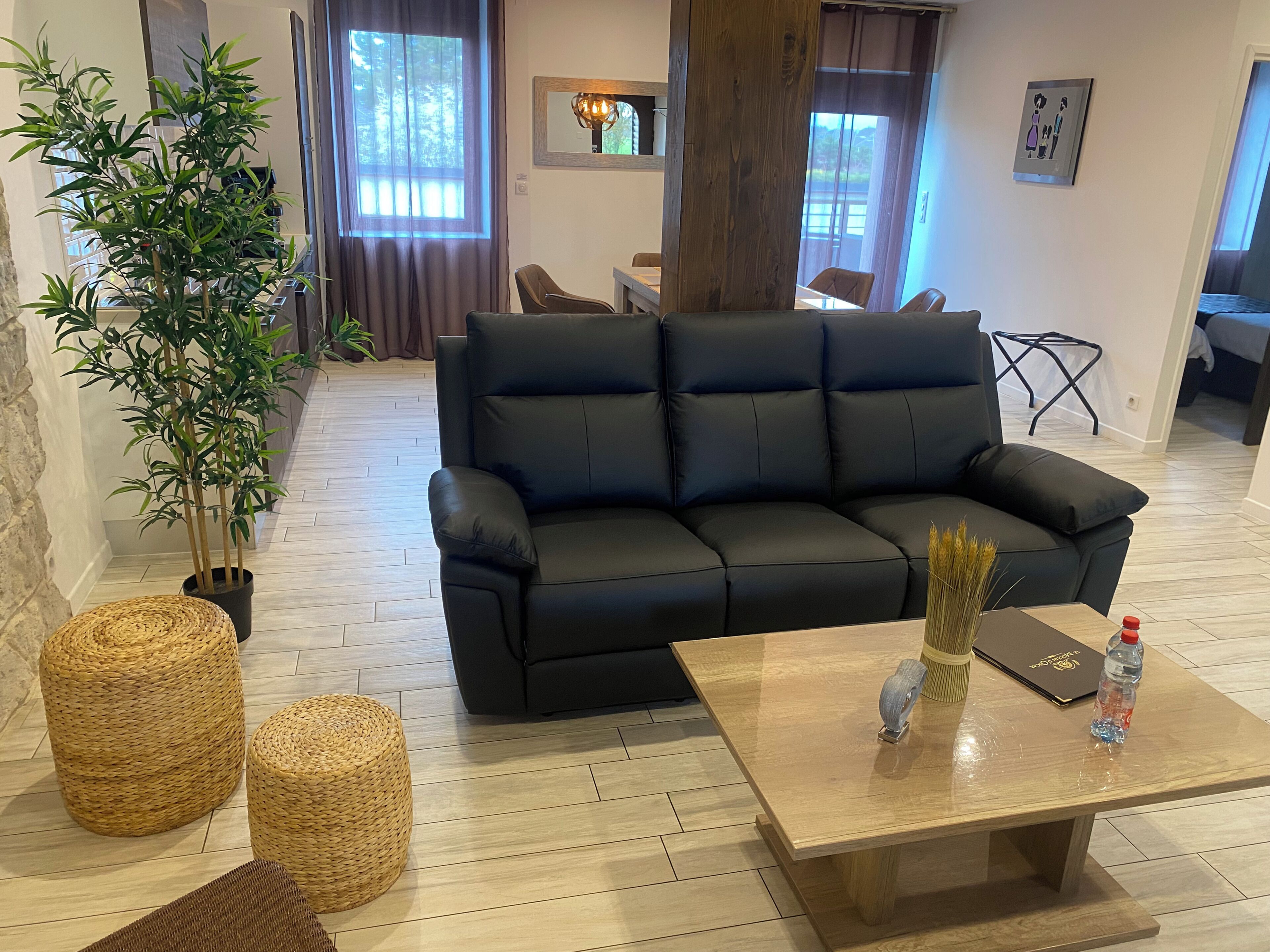 A modern living room with a black leather sofa, a wooden coffee table, wicker ottomans, and a large bamboo plant.