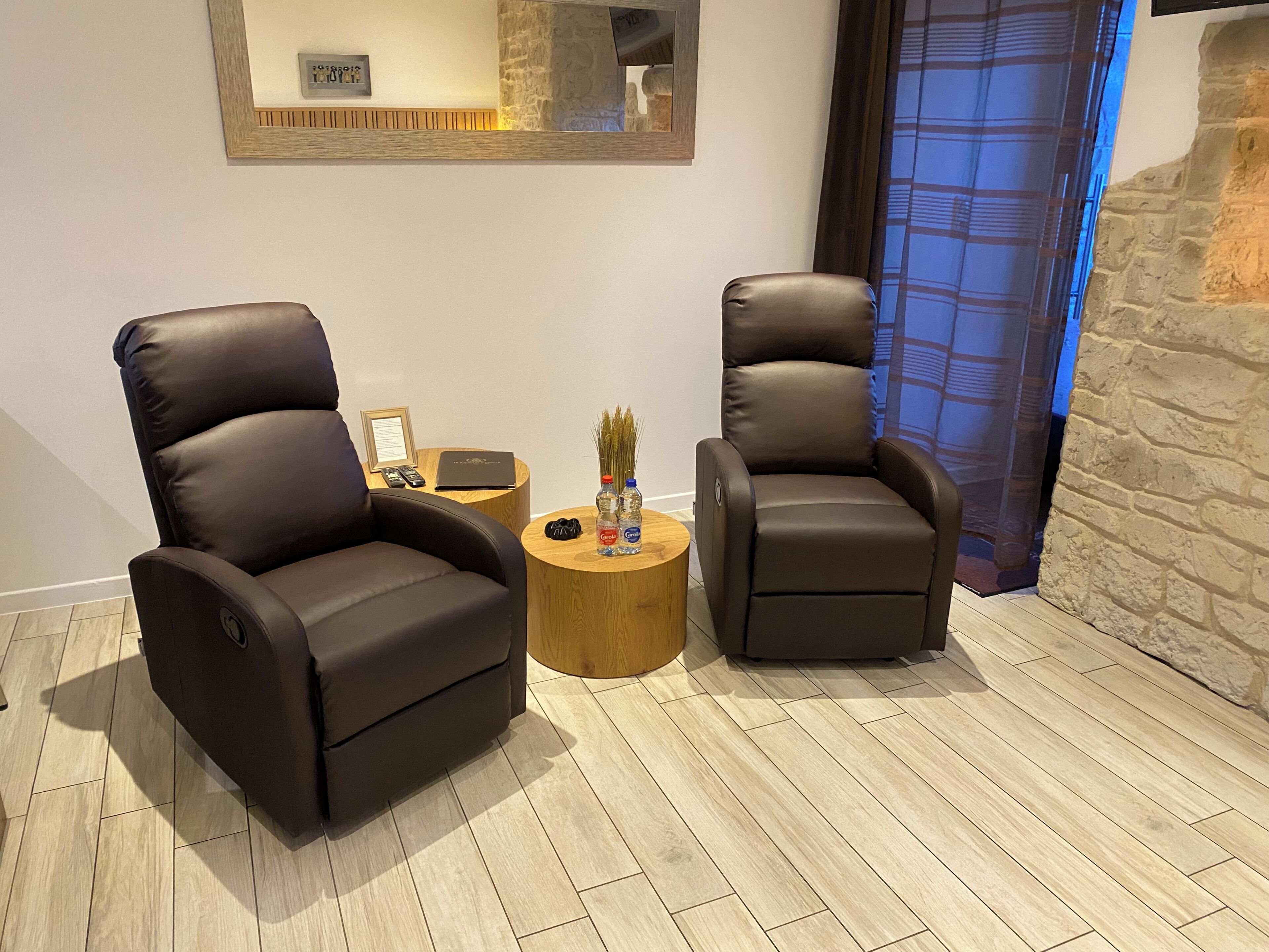Deux fauteuils inclinables en cuir, une petite table d’appoint en bois avec une bouteille et une petite plante, et un mur en pierre.
