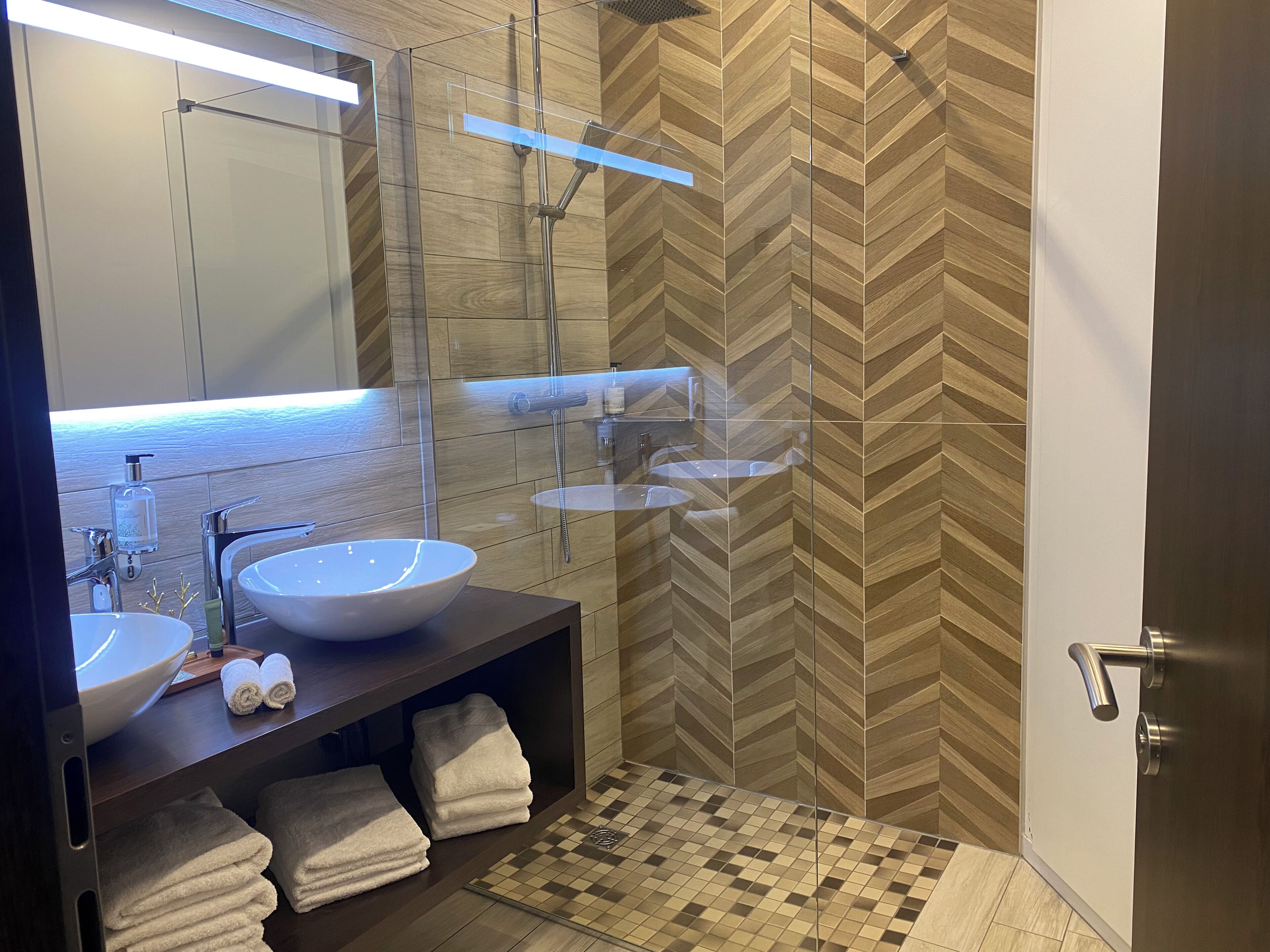 Een moderne badkamer met een grote spiegel, twee wasbakken, een douche en een wand met een herringbone-patroon.