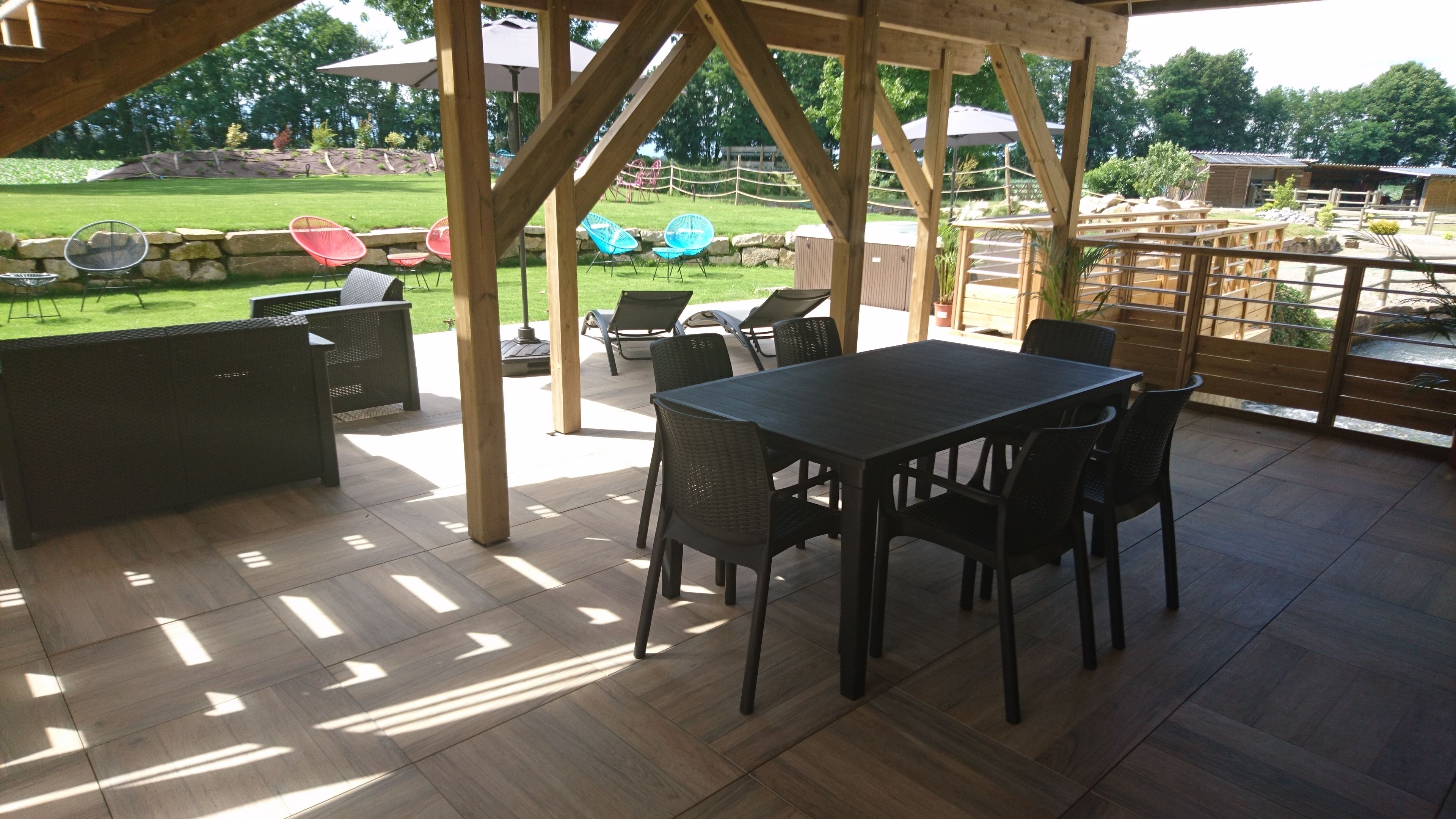 An outdoor patio with a wooden table and chairs, surrounded by a wooden fence and a view of a grassy area with chairs and umbrellas.