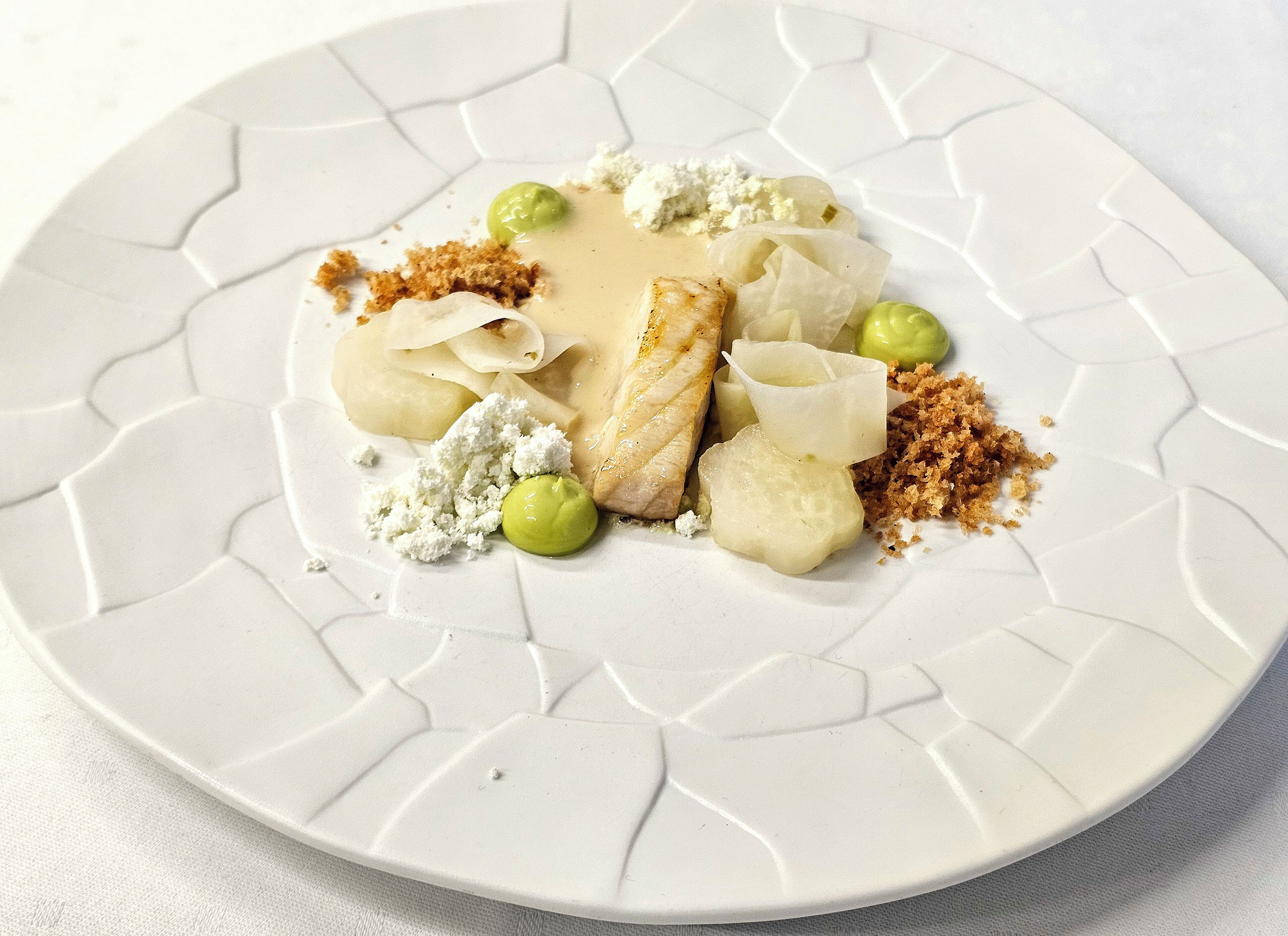 A white plate with a geometric pattern, containing a piece of fish, green peas, crumbled cheese, and a creamy sauce.