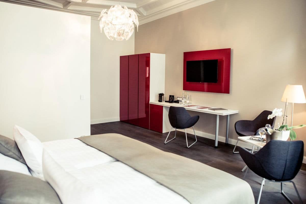 A modern hotel room with a bed, a desk, two chairs, a TV, and a chandelier.