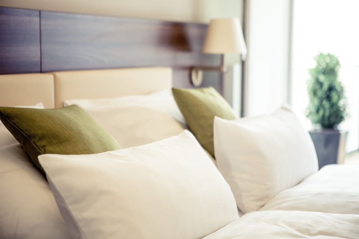 A neatly made bed with white and green pillows, a wooden headboard, and a bedside lamp.