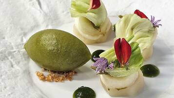 Un plato de postre con una fruta verde, posiblemente mango, y tres elementos pequeños y redondos de color blanco coronados con flores comestibles rojas y púrpuras.