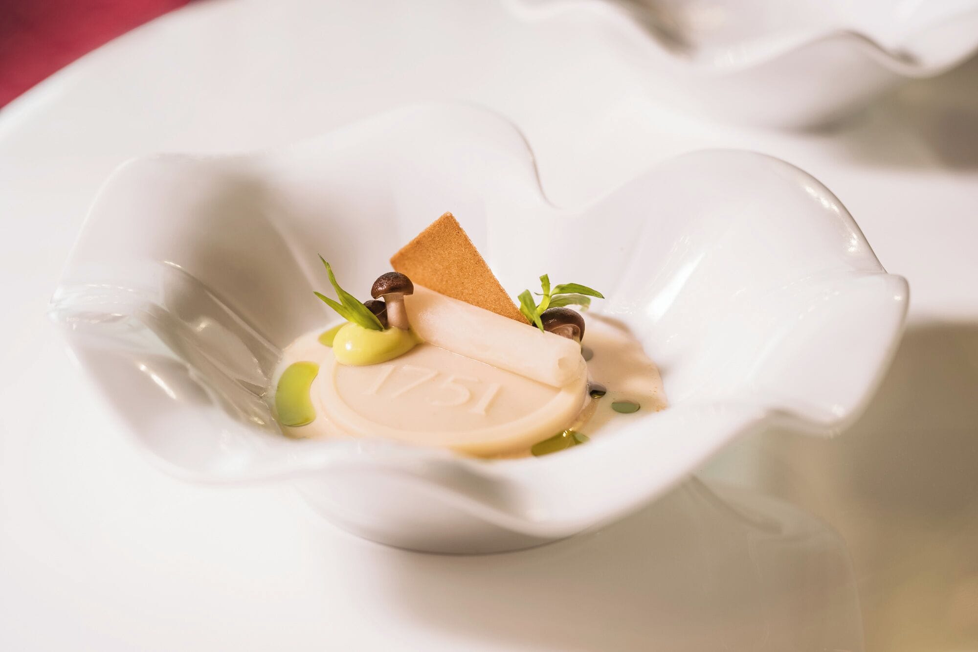 A dessert in a white, heart-shaped dish with a piece of cake, a dollop of cream, and a sprig of green herb.