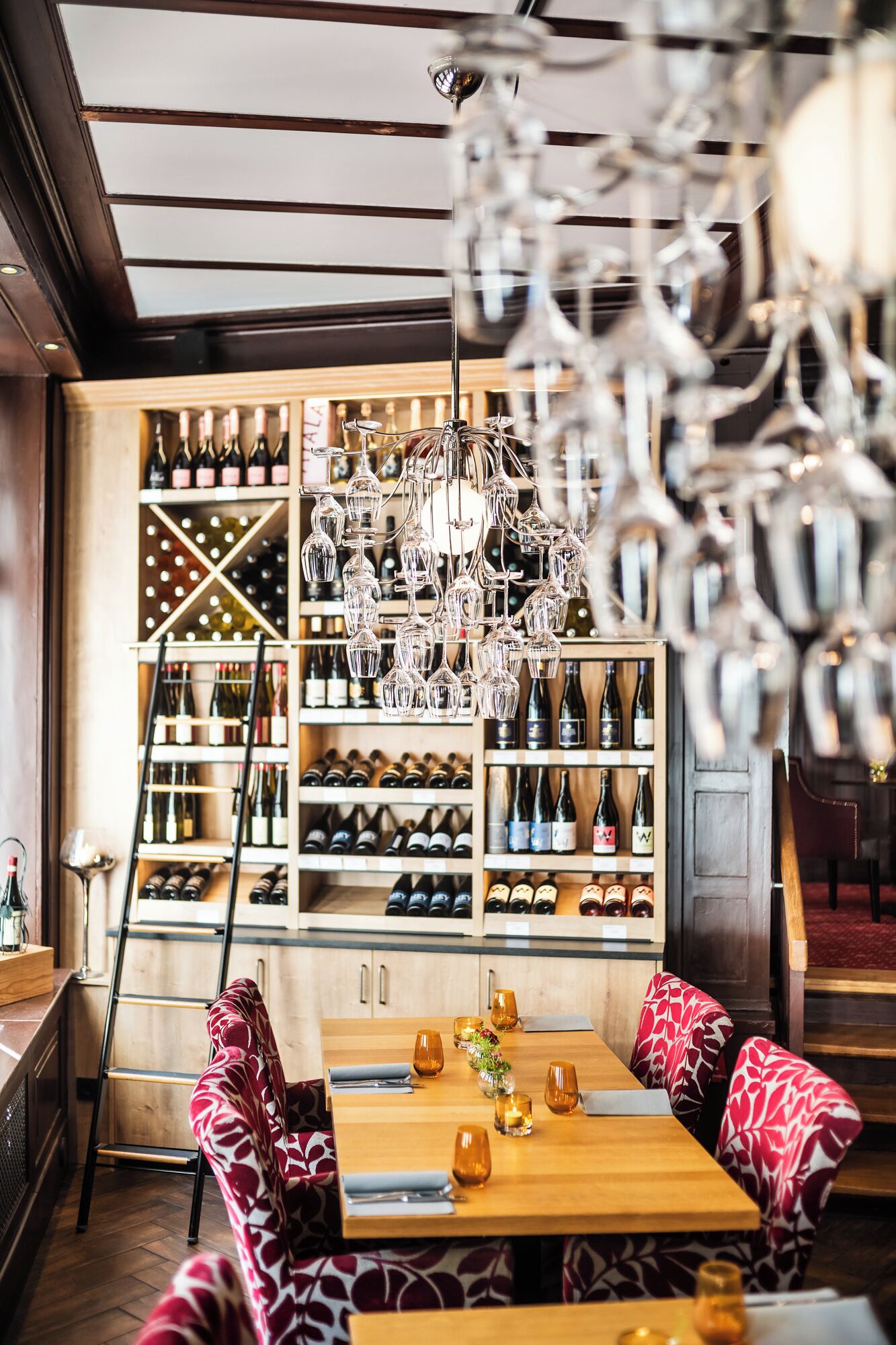 Un interior de restaurante con una mesa de madera, sillas estampadas y una estantería llena de botellas de vino.