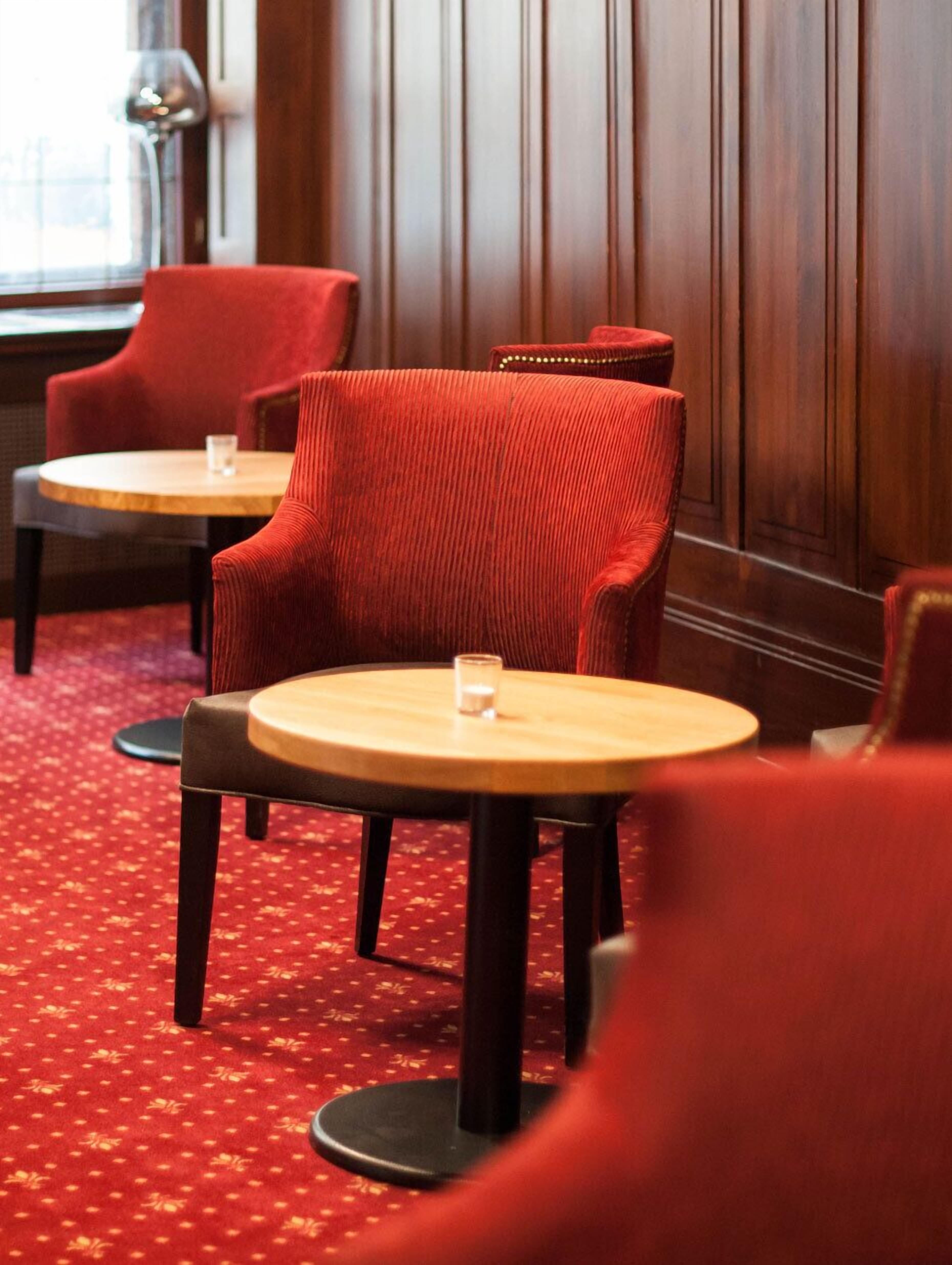 A hotel room with two red upholstered chairs and a round wooden table, each with a glass on it. The room has a red carpet and wooden paneling on the walls.