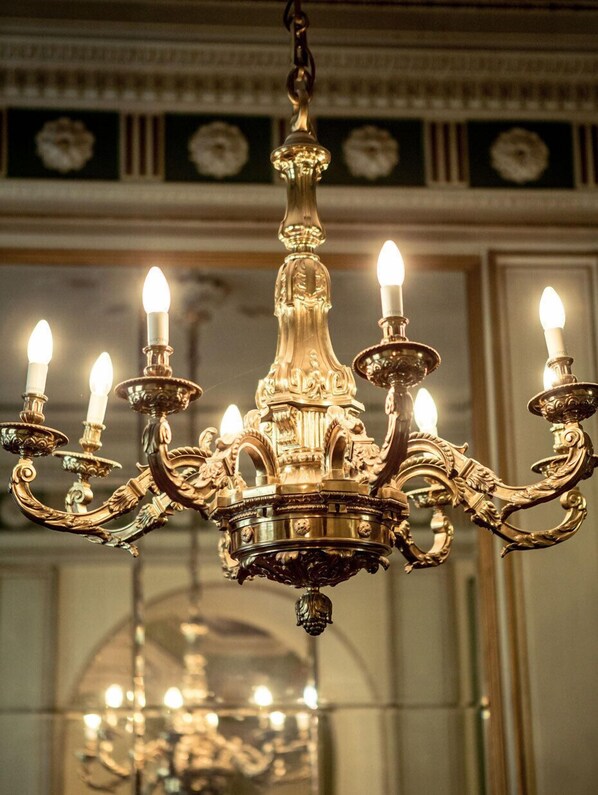 A chandelier with multiple candle-like lights, ornate design, and a mirror in the background. - Weinhaus Uhle (Schwerin)