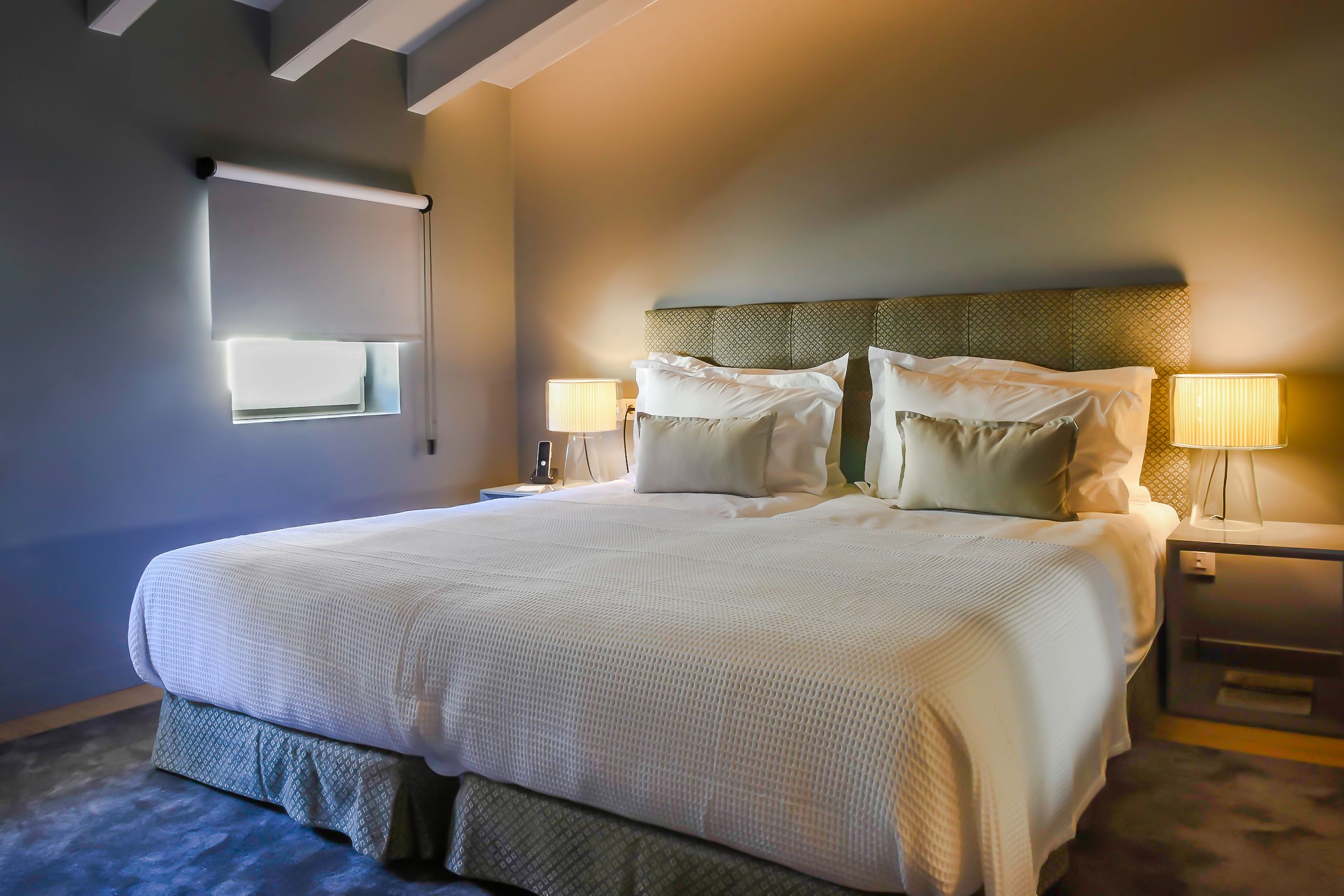 Una cama bien hecha con sábanas blancas, un cabecero con diseño geométrico y dos lámparas de noche.